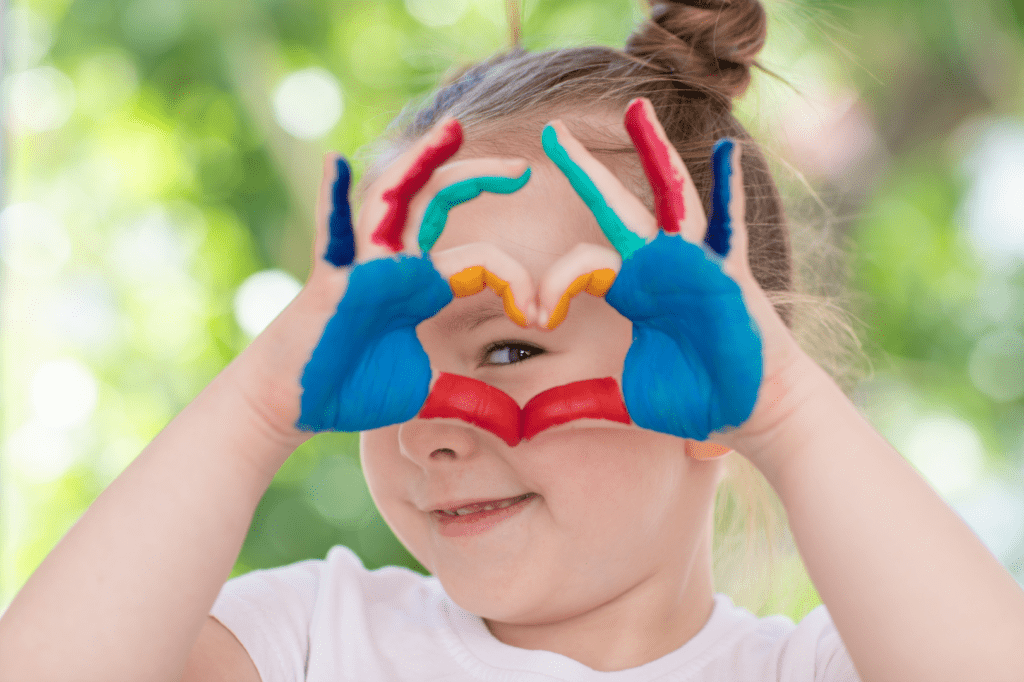 A young child peeks through a heart made from their colorful, paint-covered hands.