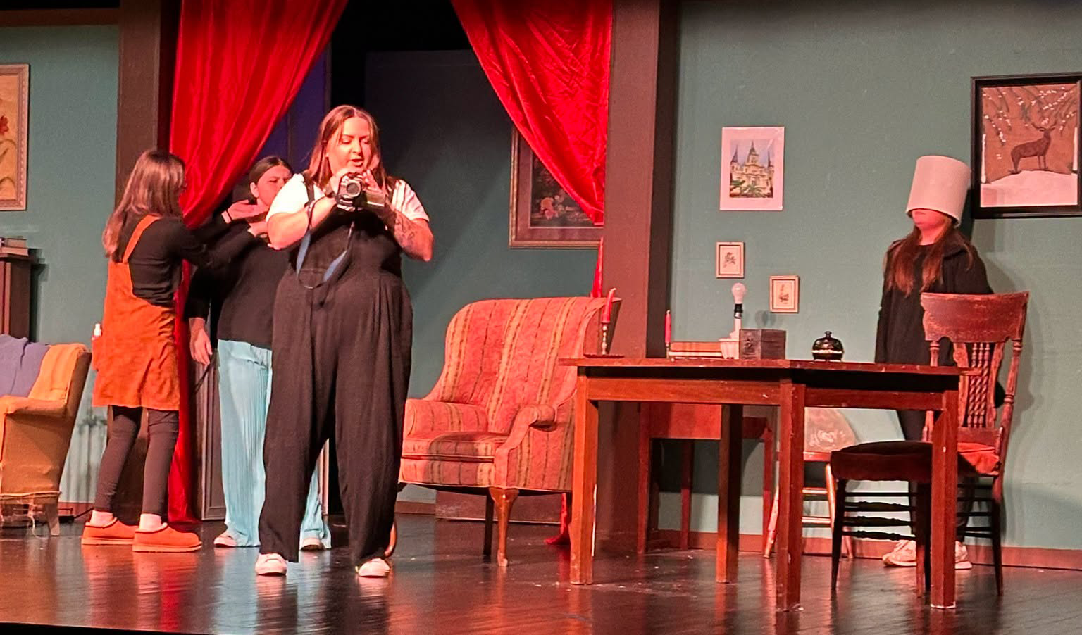 Students perform a scene on a theatre set with red curtains and stage props.